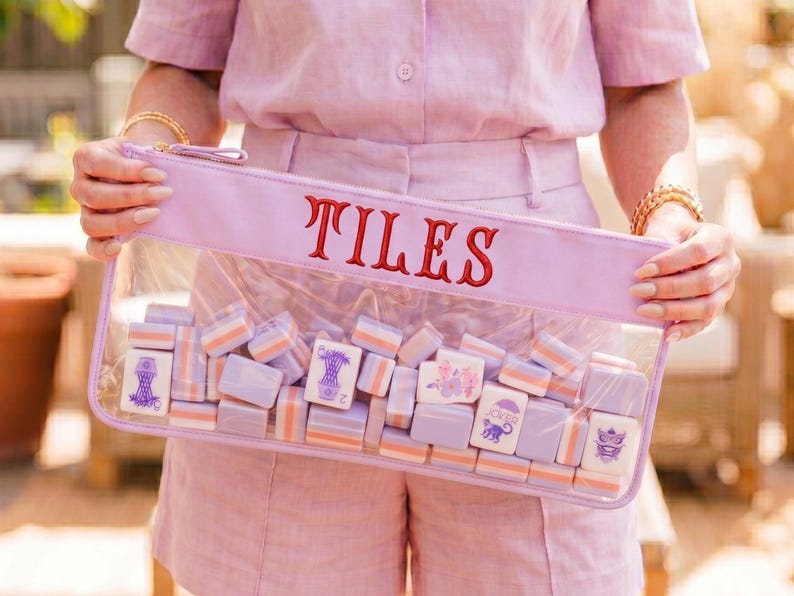 May include: A clear pouch with the word "TILES" embroidered in red. The pouch is filled with small, rectangular tiles in shades of lavender, white, and orange. The tiles feature various illustrations. The pouch is held by a person wearing a pink outfit.