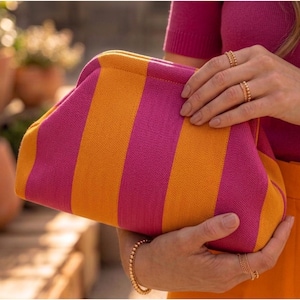 Pode incluir: Uma bolsa clutch com um padr&atilde;o vibrante de riscas verticais em tons alternados de rosa magenta brilhante e laranja dourado. A bolsa apresenta uma abertura de estrutura arredondada e uma textura de tecido de malha em todo o exterior.