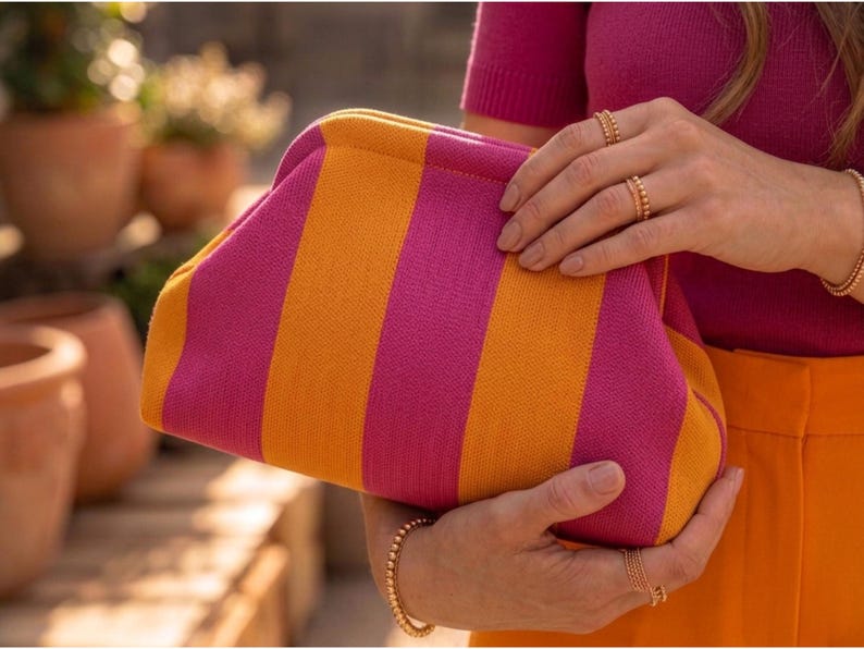 Pode incluir: Uma bolsa clutch com um padr&atilde;o vibrante de riscas verticais em tons alternados de rosa magenta brilhante e laranja dourado. A bolsa apresenta uma abertura de estrutura arredondada e uma textura de tecido de malha em todo o exterior.