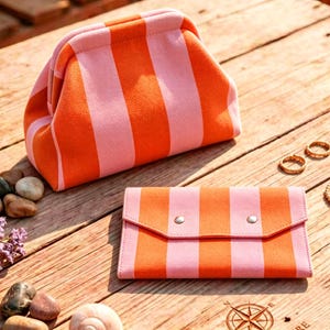 May include: A fabric pouch with a kiss-lock closure and a matching rectangular wallet, both featuring a vibrant orange and light pink vertical striped pattern. The items are displayed alongside smooth river stones and gold-toned jewelry pieces.