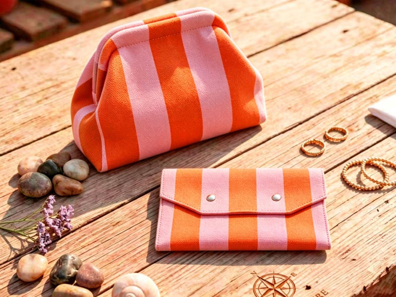 May include: A fabric pouch with a kiss-lock closure and a matching rectangular wallet, both featuring a vibrant orange and light pink vertical striped pattern. The items are displayed alongside smooth river stones and gold-toned jewelry pieces.