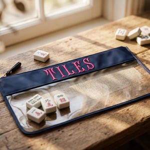 May include: A clear pouch with a navy blue top section embroidered with the word "TILES" in pink. Several Mahjong tiles are scattered around the pouch on a wooden surface. A black zip is visible.