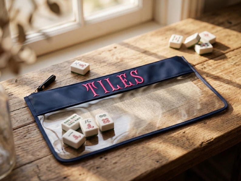 May include: A clear pouch with a navy blue top section embroidered with the word "TILES" in pink. Several Mahjong tiles are scattered around the pouch on a wooden surface. A black zip is visible.
