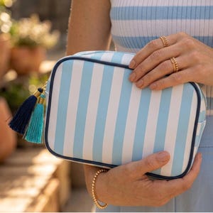 May include: A rectangular cosmetic bag with vertical light blue and white stripes. The bag has a navy blue trim and a gold zipper pull with navy and turquoise tassels. The bag is held by a person wearing gold jewelry.