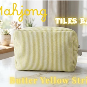 May include: A butter yellow striped Mahjong Tiles Bag with a zipper closure. The bag is rectangular and has a vertical stripe pattern. The text "Mahjong TILES BAG Butter Yellow Stripe" is displayed in yellow.