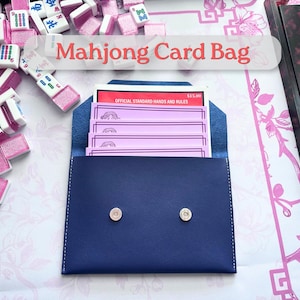 May include: A navy blue Mahjong card bag with a snap closure, containing pink cards and a red booklet. The text "Mahjong Card Bag" is displayed above the bag. Mahjong tiles are scattered around the bag.