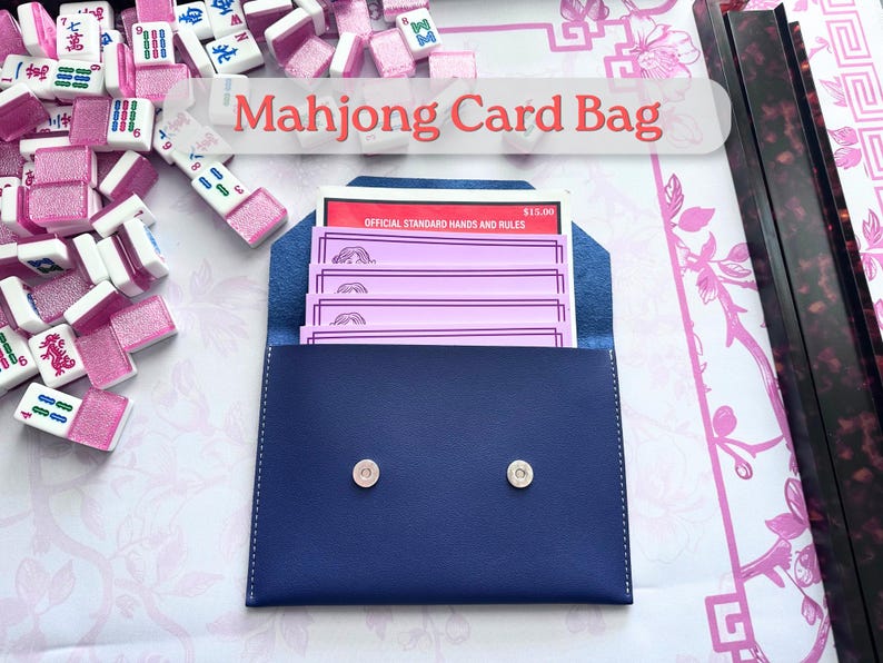 May include: A navy blue Mahjong card bag with a snap closure, containing pink cards and a red booklet. The text "Mahjong Card Bag" is displayed above the bag. Mahjong tiles are scattered around the bag.