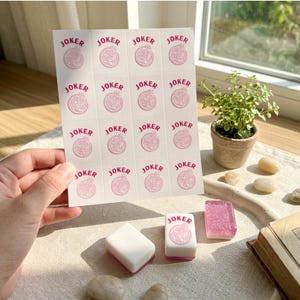 May include: A sheet of stickers with the word "JOKER" in pink, along with several Mahjong tiles. The stickers and tiles are on a table with a small potted plant and a book. The stickers have a pink design.