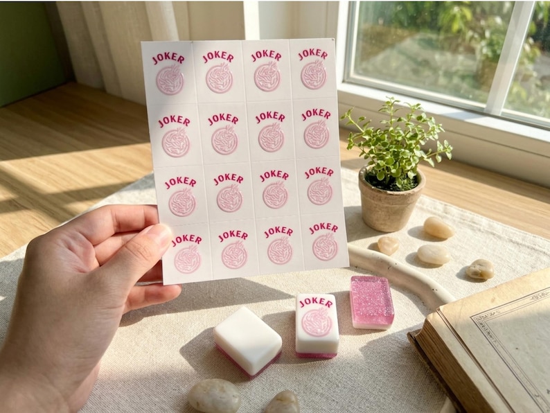 May include: A sheet of stickers with the word "JOKER" in pink, along with several Mahjong tiles. The stickers and tiles are on a table with a small potted plant and a book. The stickers have a pink design.