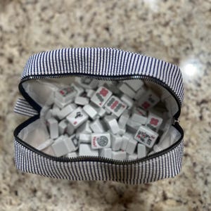 May include: A striped navy and white zippered pouch filled with white Mahjong tiles. The tiles have red and green symbols. The pouch is open, revealing the contents. The background is a speckled, light brown surface.