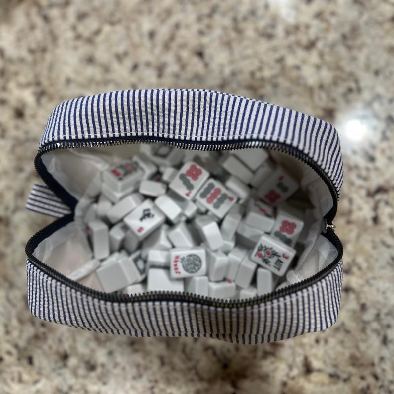 May include: A striped navy and white zippered pouch filled with white Mahjong tiles. The tiles have red and green symbols. The pouch is open, revealing the contents. The background is a speckled, light brown surface.