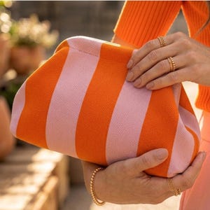 May include: A structured clutch bag featuring a vibrant knit pattern with alternating vertical stripes in bright orange and soft pastel pink. The bag has a rounded, soft-sided frame design with a concealed closure, showcasing a textured fabric finish.