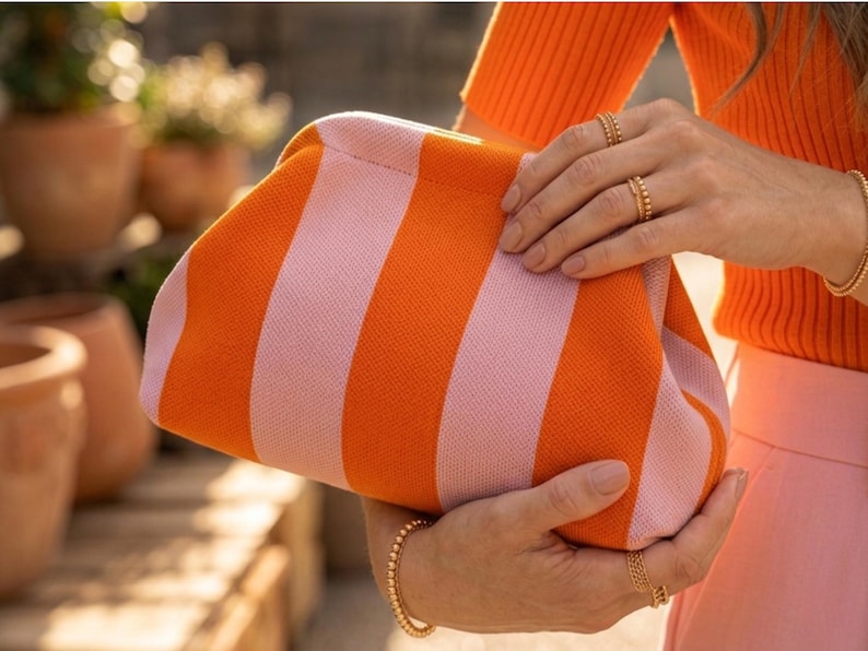 May include: A structured clutch bag featuring a vibrant knit pattern with alternating vertical stripes in bright orange and soft pastel pink. The bag has a rounded, soft-sided frame design with a concealed closure, showcasing a textured fabric finish.