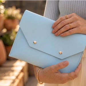 May include: A pale blue clutch bag with a flap closure and two gold-coloured button details. The bag is held by a person wearing gold jewellery. The background features terracotta pots and greenery.