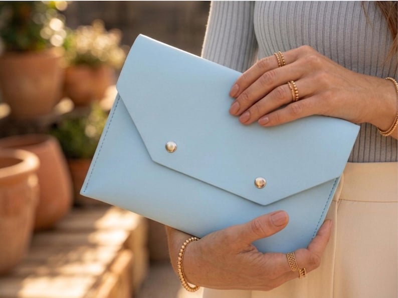 May include: A pale blue clutch bag with a flap closure and two gold-coloured button details. The bag is held by a person wearing gold jewellery. The background features terracotta pots and greenery.