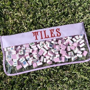 May include: A clear pouch filled with pink and white mahjong tiles. The pouch has a lavender top with the word "TILES" embroidered in red. The tiles are scattered inside the pouch, resting on a bed of green grass.