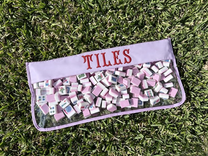 May include: A clear pouch filled with pink and white mahjong tiles. The pouch has a lavender top with the word "TILES" embroidered in red. The tiles are scattered inside the pouch, resting on a bed of green grass.