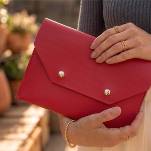 May include: A vibrant red clutch bag with a flap and two silver stud closures. The bag is held by a person wearing gold jewellery, including rings and a bracelet. The background is out of focus.