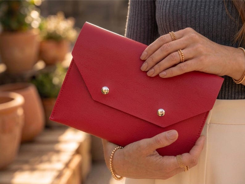 May include: A vibrant red clutch bag with a flap and two silver stud closures. The bag is held by a person wearing gold jewellery, including rings and a bracelet. The background is out of focus.