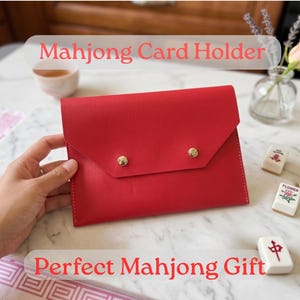 May include: A vibrant red Mahjong card holder with gold button fastenings. The image includes the text "Mahjong Card Holder" and "Perfect Mahjong Gift". Several Mahjong tiles are also visible.