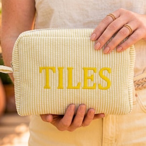 Può includere: Una pochette rettangolare con cerniera realizzata in tessuto seersucker giallo chiaro e bianco con sottili righe verticali. La parola TILES è ricamata in grassetto con lettere maiuscole giallo brillante al centro del pannello frontale.