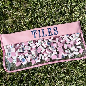 May include: A pink pouch filled with white and pink mahjong tiles, resting on green grass. The pouch has a clear front and the word "TILES" embroidered in blue across the top.