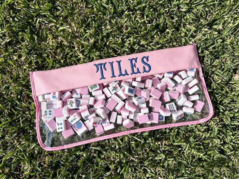 May include: A pink pouch filled with white and pink mahjong tiles, resting on green grass. The pouch has a clear front and the word "TILES" embroidered in blue across the top.