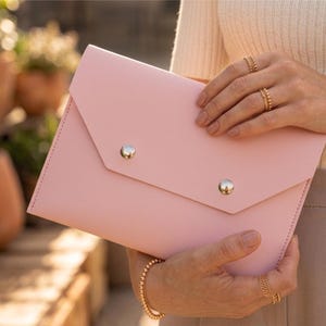 May include: A pale pink envelope clutch bag with a flap closure, secured by two silver-coloured, round studs. The bag is made of smooth material and has a stitched edge. The bag is held in the hands.