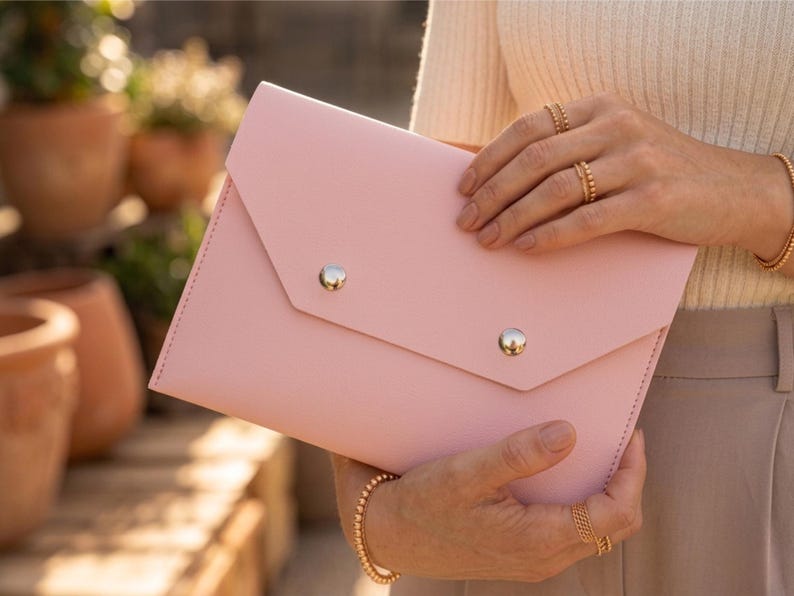 May include: A pale pink envelope clutch bag with a flap closure, secured by two silver-coloured, round studs. The bag is made of smooth material and has a stitched edge. The bag is held in the hands.