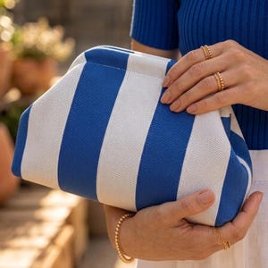 May include: A clutch bag featuring a bold pattern of vertical royal blue and white stripes. The bag has a structured, rounded frame design with a textured fabric exterior. The clutch is held in place, showcasing its clean lines and color contrast.