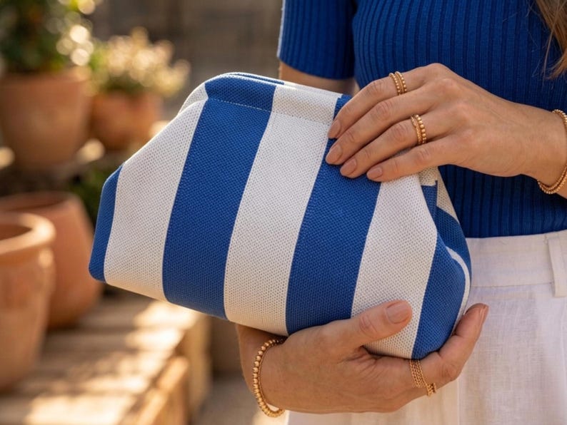 May include: A clutch bag featuring a bold pattern of vertical royal blue and white stripes. The bag has a structured, rounded frame design with a textured fabric exterior. The clutch is held in place, showcasing its clean lines and color contrast.
