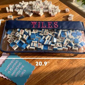May include: A clear pouch filled with white and blue mahjong tiles, with the word "TILES" embroidered in pink. The pouch is 53.1 cm long and 23.1 cm high. A few tiles are scattered around the pouch. A card reads "HAPPINESS IS PLAYING MAHJONG WITH THE GIRLS."