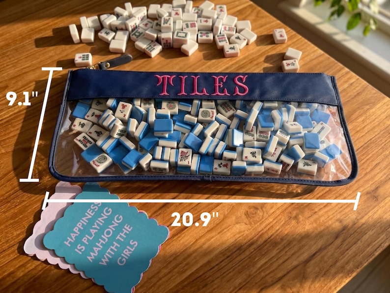May include: A clear pouch filled with white and blue mahjong tiles, with the word "TILES" embroidered in pink. The pouch is 53.1 cm long and 23.1 cm high. A few tiles are scattered around the pouch. A card reads "HAPPINESS IS PLAYING MAHJONG WITH THE GIRLS."