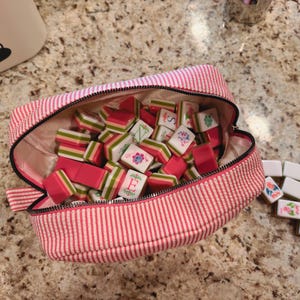 May include: A pink and white striped zippered pouch filled with a collection of colorful Mahjong tiles. The tiles are primarily red, white, and green, with various symbols and letters. The pouch is open, revealing the contents.