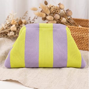 May include: A structured clutch bag featuring a knit texture with vertical stripes in alternating shades of bright lime green and soft lavender. The bag has a curved top frame design and a wide, trapezoidal shape with rounded corners.