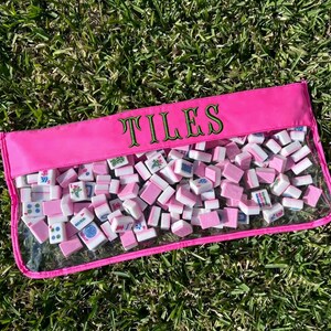 May include: A pink rectangular pouch filled with white and pink mahjong tiles. The pouch has a clear front and the word "TILES" embroidered in green. The tiles are scattered inside the pouch, resting on green grass.