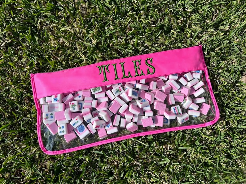 May include: A pink rectangular pouch filled with white and pink mahjong tiles. The pouch has a clear front and the word "TILES" embroidered in green. The tiles are scattered inside the pouch, resting on green grass.