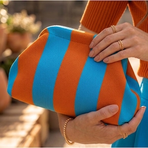 May include: A clutch bag featuring a vibrant striped pattern in alternating bright orange and sky blue colors. The bag has a soft, textured knit fabric construction with a rounded, structured frame opening at the top for easy access to contents.