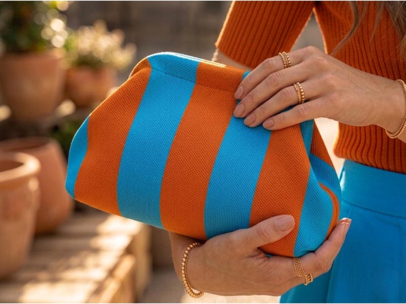 May include: A clutch bag featuring a vibrant striped pattern in alternating bright orange and sky blue colors. The bag has a soft, textured knit fabric construction with a rounded, structured frame opening at the top for easy access to contents.