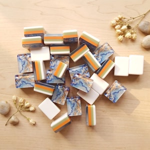 May include: A collection of rectangular mahjong tiles featuring three distinct designs: solid white, a layered pattern of orange, green, and white stripes, and a blue and gold marbled swirl pattern. The tiles are scattered across a light wood surface.
