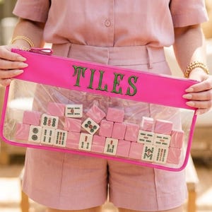May include: A bright pink clear pouch with the word "TILES" in green lettering. The pouch contains pink mahjong tiles. The person in the image is wearing a pink outfit.