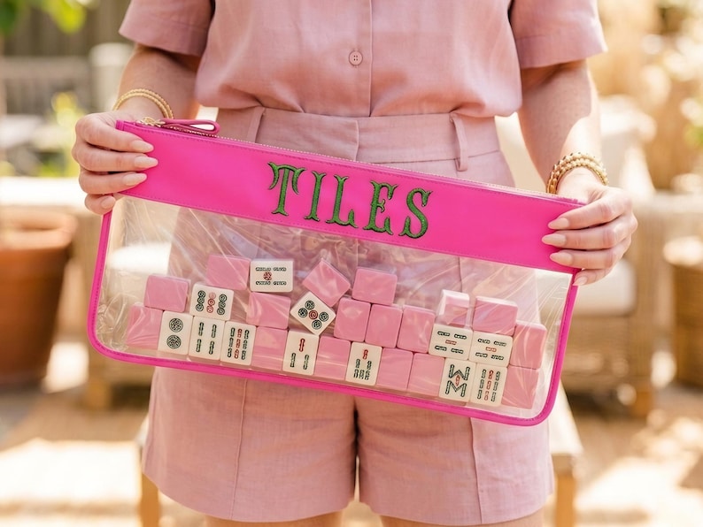 May include: A bright pink clear pouch with the word "TILES" in green lettering. The pouch contains pink mahjong tiles. The person in the image is wearing a pink outfit.