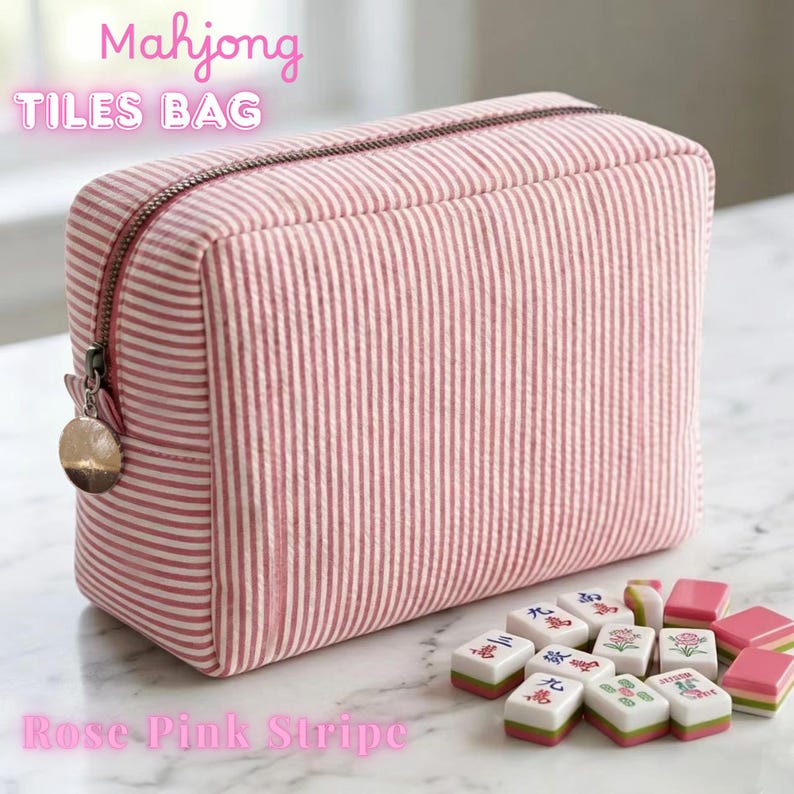May include: Rose pink striped Mahjong tiles bag with a zipper closure and a metal charm. The bag is rectangular and has a vertical stripe pattern. Mahjong tiles are arranged next to the bag.