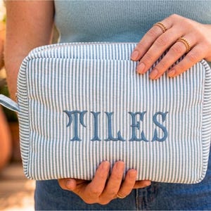 Könnte beinhalten: Eine rechteckige Kosmetiktasche mit blau-weißen Streifen und dem Wort "TILES" in Blau gestickt. Die Tasche hat einen Reißverschluss und wird von einer Person gehalten.