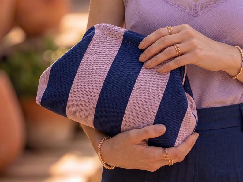 Pode incluir: Uma bolsa clutch com um padr&atilde;o de riscas verticais em azul-marinho e rosa claro. A bolsa tem uma estrutura superior arredondada e um exterior em tecido texturizado. &Eacute; segurada por duas m&atilde;os adornadas com an&eacute;is de tom dourado.