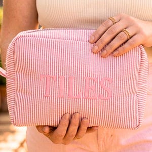 May include: Pink and white striped cosmetic bag with the word "TILES" embroidered in pink. The bag has a rectangular shape with a zipper closure and a small loop on the side.