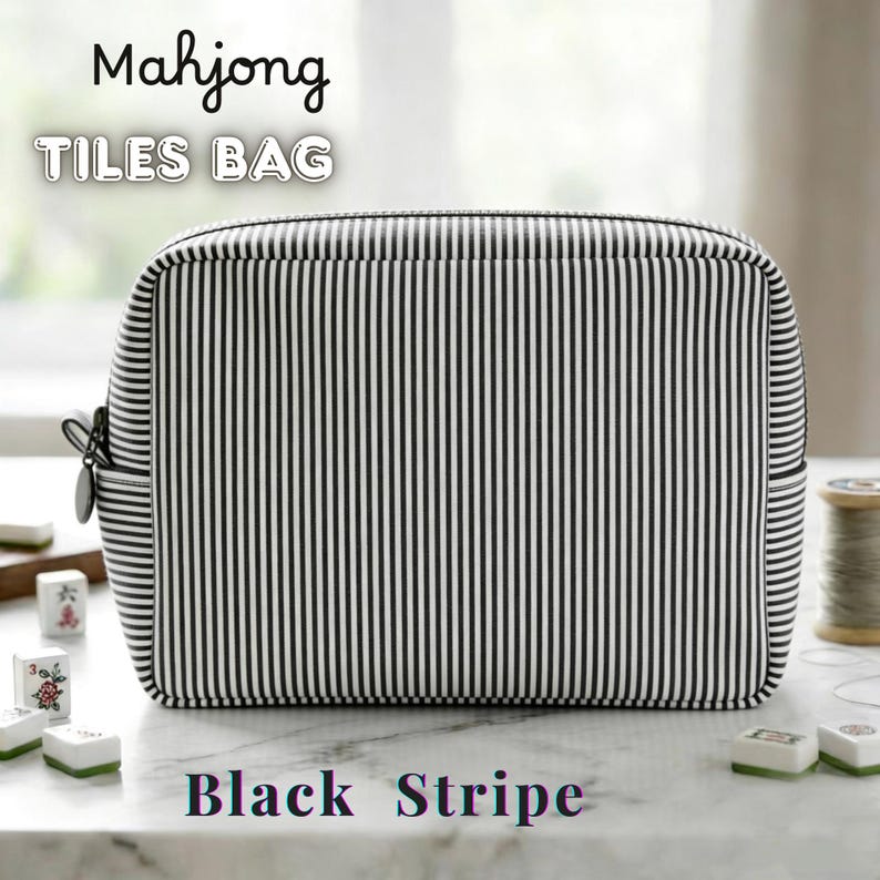 May include: A rectangular black and white striped bag with the text "Mahjong TILES BAG" at the top and "Black Stripe" at the bottom. The bag has a zipper closure and is sitting on a white surface. Mahjong tiles are visible.