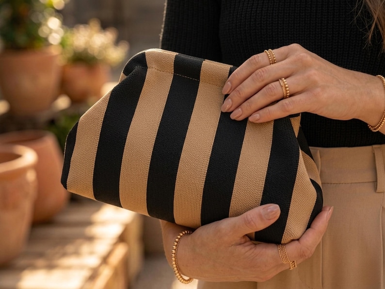 May include: A structured clutch bag featuring a bold vertical striped pattern in alternating black and tan colors. The bag has a soft, textured fabric exterior with a curved top frame closure, held securely by two hands adorned with gold rings.