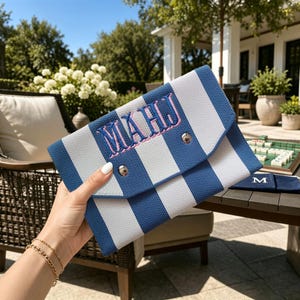 May include: A blue and white striped envelope-style clutch bag featuring the embroidered letters "MAHJ" in pink with a blue outline. The bag has a triangular flap secured by a silver-toned metal snap button closure on the front exterior.