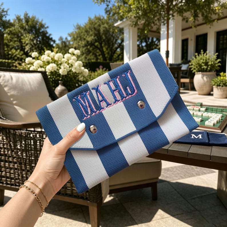 May include: A blue and white striped envelope-style clutch bag featuring the embroidered letters "MAHJ" in pink with a blue outline. The bag has a triangular flap secured by a silver-toned metal snap button closure on the front exterior.